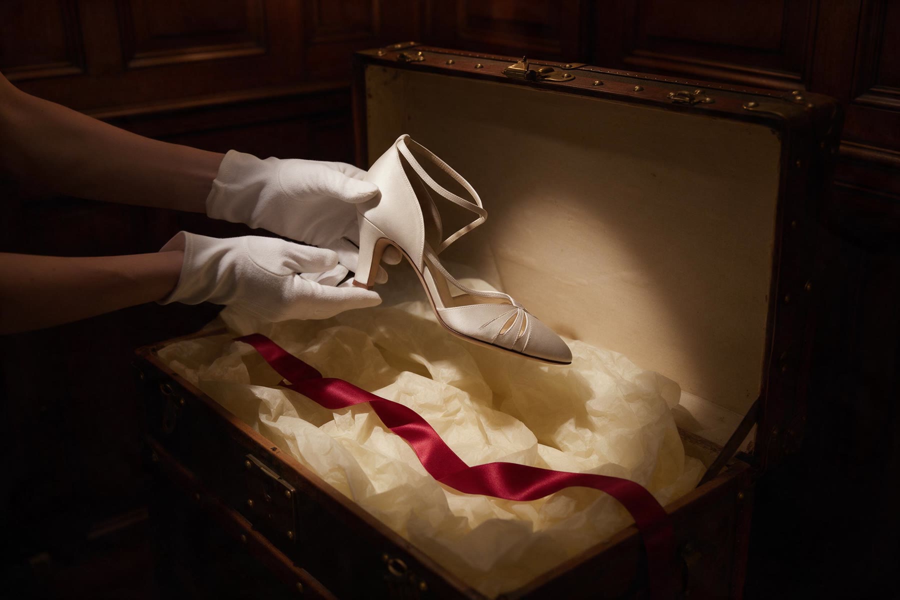 White-gloved hands lifting a shoe from a tissue-lined steamer trunk